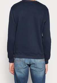 Marineblauer Pullover mit Rundhalsausschnitt, gerippten Bündchen und Saum, kombiniert mit hellblauen Jeans, die schräg angesetzte Gesäßtaschen und sichtbare Nähte haben.