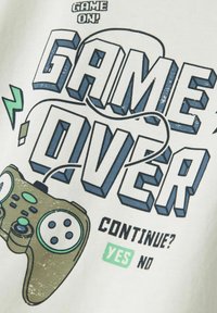 Tessuto bianco con testo audace blu e verde "GAME OVER" e un'immagine di un controller di gioco marrone. Il testo include "CONTINUARE? SÌ NO."