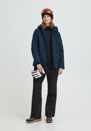 Kobieta w ciemnej kurtce narciarskiej, czarnych spodniach narciarskich i goglach narciarskich trzymająca białe i czarne rękawice narciarskie na jednolitym tle.