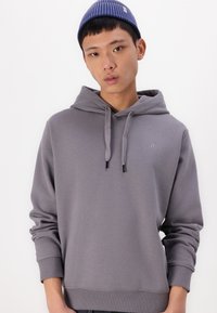 Sudadera gris con cordón y bolsillo frontal tipo canguro. Textura suave, puños acanalados y un pequeño logo en el pecho. Usada con un gorro azul.
