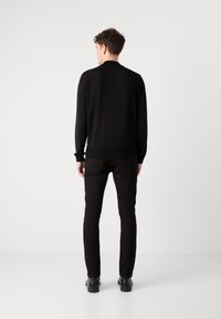 Pull noir à manches longues avec un col rond et des poignets côtelés, associé à un pantalon noir slim et des chaussures noires, vu de dos.