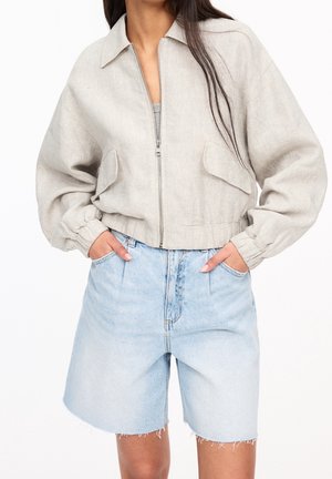 Femme portant une veste beige zippée avec des poignets élastiques et un short en jean bleu clair taille haute aux ourlets effilochés. Mains dans les poches.