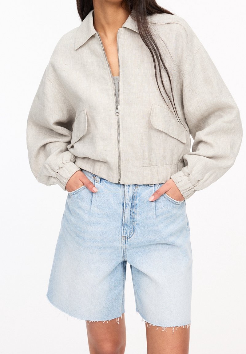 Femme portant une veste beige zippée avec des poignets élastiques et un short en jean bleu clair taille haute aux ourlets effilochés. Mains dans les poches.