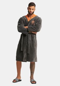 Houtskoolgrijze badjas met een oranje capuchon, een strikceintuur, twee voorzakken en een gestructureerde stof. Model draagt schuifsandalen met een patroonontwerp.