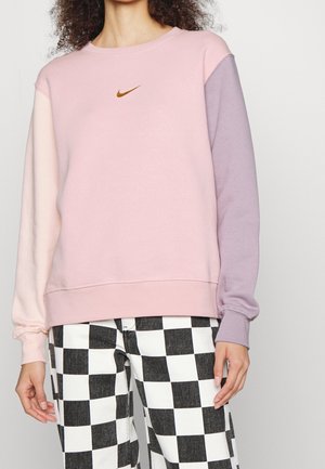 Osoba ubrana w różowo-lawendową bluzę z małym brązowym logo Nike oraz czarno-białe spodnie w kratę.