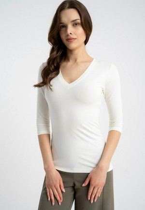 Młoda kobieta o długich brązowych włosach, ubrana w biały top z dekoltem w serek i rękawami do łokcia oraz oliwkowe spodnie, stojąca na jednolitym tle.