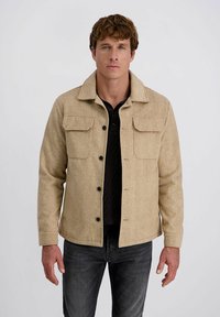 Man in een beige herringbone jack met zwarte knopen over een zwart shirt en donkere jeans, staand tegen een effen witte achtergrond.