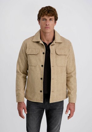 Homme portant une veste en chevron beige avec des boutons noirs sur une chemise noire et un jean foncé, se tenant devant un fond blanc uni.