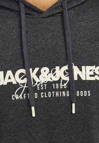 Donkergrijze hoodie met trekkoorden, voorzien van een groot wit logo en tekstontwerp. Stof lijkt zacht met een gestructureerde afwerking.