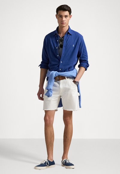 Blauwe overhemd met knopen over een gestreept T-shirt, witte shorts met rafelige randen en blauwe sneakers. Accessoires zijn onder andere een zonnebril en een horloge.