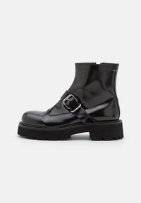Botas negras hasta el tobillo con un acabado brillante, que cuentan con una correa con hebilla, punta redondeada y una suela gruesa y texturizada para mayor agarre. Textura de cuero suave.