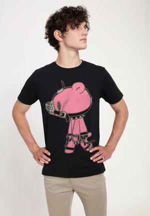 Ung mand med krøllet hår iført en sort t-shirt med en lyserød bamse tegneserie i sele og mundkurv, stående med hænderne på hofterne.