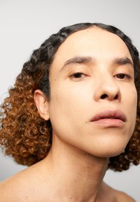 Primo piano di una persona con capelli ricci di lunghezza media, con radici più scure e punte più chiare. La pelle è liscia con alcune lentiggini, tonalità neutre.