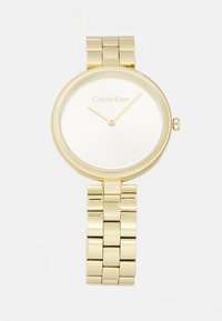 Montre-bracelet en métal doré avec un cadran rond lisse, une lunette rainurée, un cadran blanc et des index d'heure simples. Elle est dotée d'un bracelet à maillons.