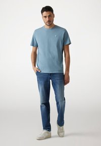 T-shirt in cotone azzurro chiaro con scollo rotondo e piccolo logo sul petto, abbinata a jeans blu scoloriti e sneakers bianche.