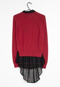 Haut rouge à manches longues avec un dos à col noir, présentant une couche inférieure en dentelle à carreaux noirs et un ourlet froncé. Suspendu à un crochet.