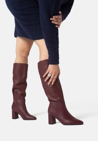 Stivali in pelle bordeaux al ginocchio con tacco a blocco e chiusura con zip, caratterizzati da una superficie liscia e una punta appuntita.