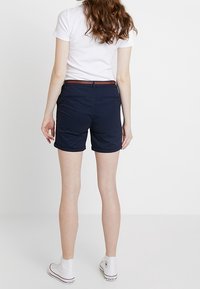 Marinblå shorts i lätt tyg med en åtsittande design och en brun bältesdetalj. Kombineras med vita sneakers.