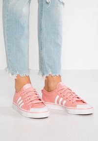 Le sneakers di tela rosa presentano strisce laterali bianche, suola in gomma bianca e lacci coordinati, abbinate a jeans in denim azzurro chiaro sfilacciati.