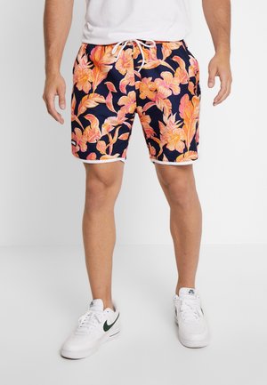 Pantaloni da bagno navy con un vivace motivo floreale arancione, rifinitura bianca e vita regolabile con cordino. Il modello indossa sneakers bianche con dettagli verdi.