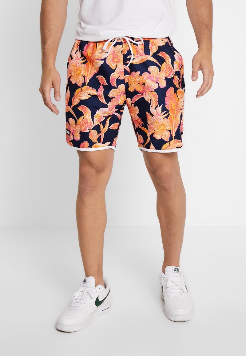 Pantaloni da bagno navy con un vivace motivo floreale arancione, rifinitura bianca e vita regolabile con cordino. Il modello indossa sneakers bianche con dettagli verdi.