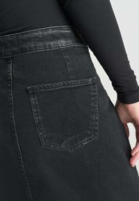 Zwarte denim rok met een hoge tailleband en een geborduurd achterzak met een gebogen patroon. Soepele textuur en nauwsluitende pasvorm.