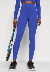 Plave tajice visokog struka izrađene od rastezljivog materijala, s glatkom teksturom i prilagođenim dizajnom, uparenim s crnim sportskim cipelama i palicom.