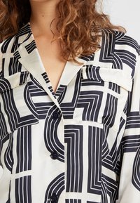 Chemise blanche à manches longues ornée d'un motif géométrique noir, avec deux poches poitrine et une texture soyeuse, dotée d'un col à rabats.