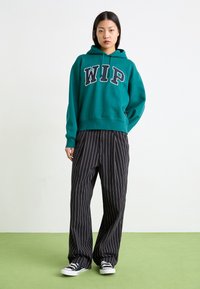 Felpa color turchese con la scritta "WIP" in bianco, abbinata a pantaloni a gamba larga neri a righe e sneakers nere basse su un pavimento verde.