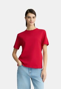 T-shirt in cotone rosso con maniche corte e scollatura rotonda, vestibilità rilassata, indossata con jeans in denim blu chiaro. Texture liscia, senza motivi visibili.