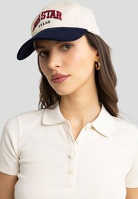 Casquette beige avec visière marine, comportant le texte brodé rouge "BIG STAR JEANS." Vue rapprochée d'une femme portant un polo à côtes léger.