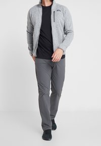 Grå fleecejacka med hög krage, framficka med dragkedja, svart t-shirt under, grå byxor och svarta joggingskor.