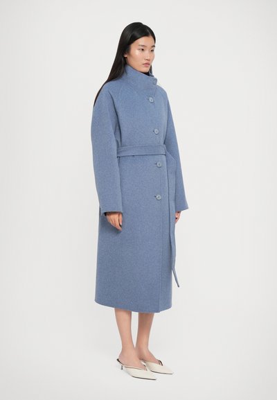 Femme portant un manteau en laine bleu ceinturé et boutonné avec un col montant, se tenant devant un fond blanc uni, en talons crème pointus.