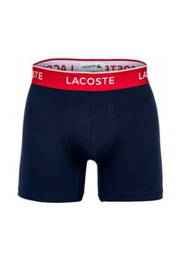 Boxer in cotone blu navy con elastico rosso in vita, caratterizzati da un marchio "LACOSTE" bianco e un logo. Design aderente con dettagli di cucitura.