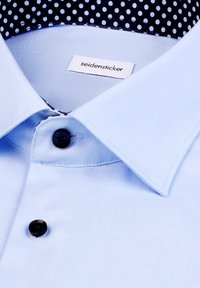 Camicia azzurro chiaro con bottoni neri, colletto appuntito e collo interno a pois blu navy. L'etichetta riporta "seidensticker". Texture liscia.