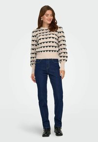 Suéter de punto con patrón de corazones en beige y negro, con dobladillo acanalado y mangas abullonadas; combinado con jeans azul oscuro y botas negras.