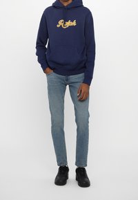 Sudadera azul marino con bordado amarillo "Ralph", bolsillo frontal, combinada con jeans azul claro y zapatillas negras. Diseño simple, ajuste casual.