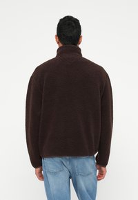 Jachetă din fleece marron cu guler înalt și croială relaxată. Suprafață texturată și umeri căzuți; asortată cu blugi din denim de culoare bleu deschis.