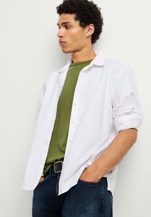 Jonge man met krullend haar, een wit overhemd over een olijfgroen T-shirt en donkere spijkerbroek, handen in de zakken, kijkt naar de zijkant.
