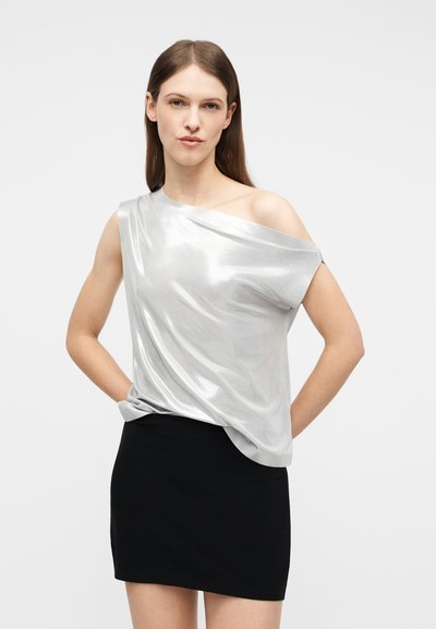 Γυναίκα με μακρυά καστανά μαλλιά, φορώντας ένα λαμπερό ασημένιο τοπ ενός ώμου και μια μαύρη μίνι φούστα, στέκεται με τα χέρια στο πίσω μέρος.