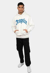 Mand iført hvid hættetrøje med blå "Dropsize" tekst, sorte cargo-bukser og hvide sneakers, stående med hænderne i hættetrøjens lommer.