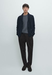 Dark navy ribbed cardigan over grey top, paired with dark brown trousers and black shoes. Simple design with button details, textured knit.
