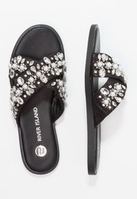Svarta slidesandaler med en strukturerad rem prydd med klara och vita pärlor, utrustade med en platt svart sula och logotyp.