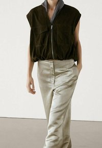 Femme portant un gilet sans manches marron foncé zippé sur une chemise rayée et un pantalon taille haute beige clair, debout avec une main dans la poche.