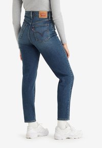 Femme portant un jean taille haute bleu avec poches arrière et des baskets blanches, debout avec une jambe légèrement pliée contre un fond blanc.