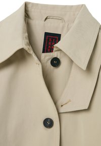 Beige trenchcoat met een klassieke kraag, voorzien van zwarte knopen en een label met rode tekst. Gladde textuur en minimalistisch ontwerp.