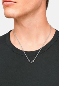 Silberne Kettenhalskette mit einem schwarzen Anhänger, der metallische Akzente aufweist; getragen über einem dunklen T-Shirt, das die Handwerkskunst und Designdetails zur Schau stellt.