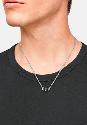 Silberne Kettenhalskette mit einem schwarzen Anhänger, der metallische Akzente aufweist; getragen über einem dunklen T-Shirt, das die Handwerkskunst und Designdetails zur Schau stellt.
