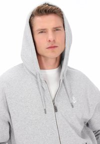 Sweat à capuche gris zippé avec cordon de serrage, présentant un petit logo ancre blanche sur la poitrine gauche et des fermetures éclair argentées.