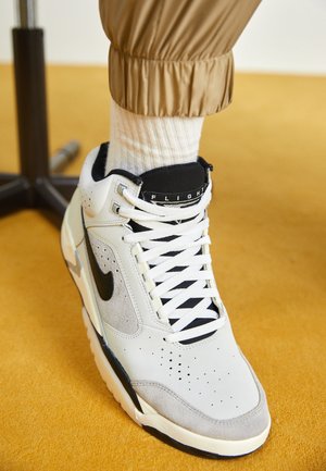 Weißer und grauer Nike-Sneaker mit schwarzem Swoosh-Logo, getragen mit weißen, gerippten Socken und beigefarbenen, oberhalb des Knöchels umgeschlagenen Hosen auf gelbem Boden.